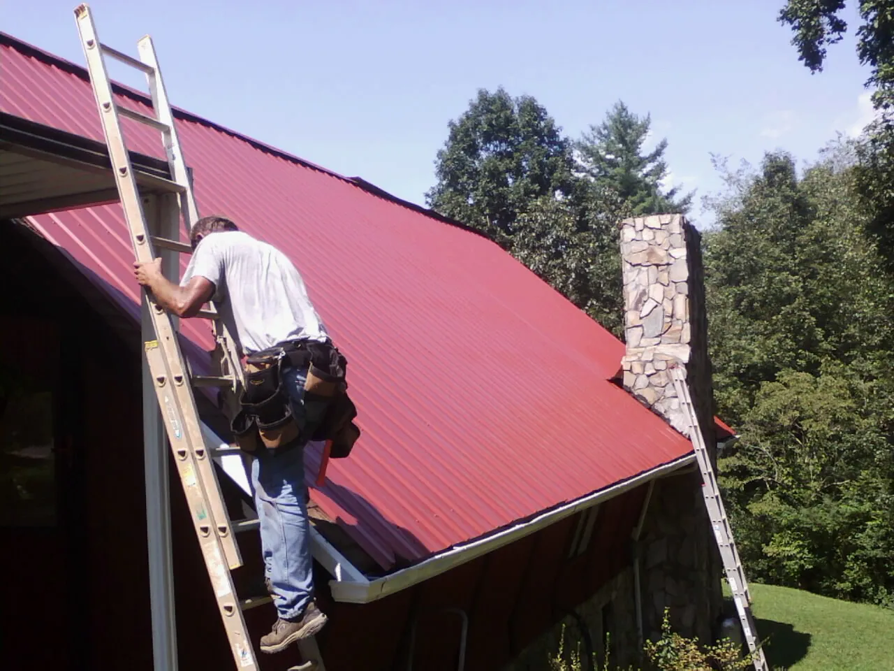 Roofer on a ladder beside a red metal roof and stone chimney — Soffit & Fascia Repair by Roofing Brothers in Alvo